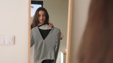 Genç bir esmer kadın hangi elbiseyi giyeceğini düşünüyor, bir beden mi yoksa kırmızı bir elbise mi, aynada kendine seçenekler sunuyor..