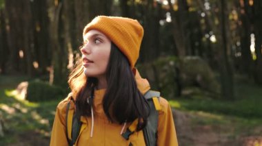 Sarı ceketli ve bereli genç bir kadın ormanda yürüyor. Doğayı keşfederken sağa sola bakıyor..