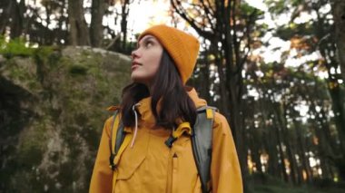 Sarı ceketli ve bereli bir kadın, sırt çantalı, bereketli bir ormanı keşfederken düşünceli bir şekilde uzaklara bakar..