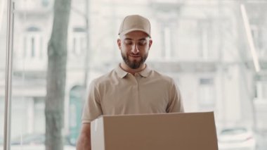 Günlük kıyafetli gülümseyen bir teslimatçının portresi. Elinde bir karton kutu, kendine güvenen bir şekilde bir ofis ortamında duruyor..