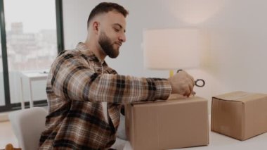 Ekose gömlekli neşeli bir adam evinde makas kullanarak yeni bir teslimat kutusunu açıyor. Ortam çok rahat ve paketleri açmanın heyecanını ve beklentisini anlatıyor..