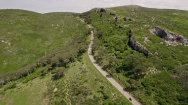 Yukarıdaki manzara, yeşil dağlık arazide, dolambaçlı bir yolda gezinen motosikletçileri yakalıyor, macera ve özgürlük hissi veriyor..