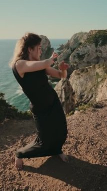 Siyah elbiseli genç bir kadın okyanusa bakan kayalık bir uçurumda zarif bir şekilde dans ediyor, özgürlük ve zerafet anı yakalıyor..