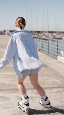 Gündelik kıyafetlerle, güneşli bir iskelede deniz manzaralı paten kayan genç bir kadının arka görüntüsü..