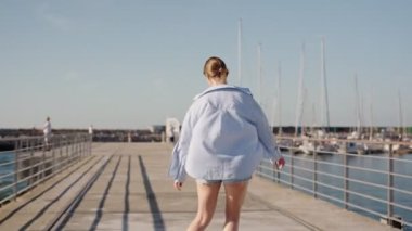 Kaygısız genç bir kadının güneşli bir iskelede paten yapıp dans edişinin arka görüntüsü. Kot şort ve açık renk gömlek giyiyor..