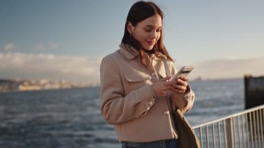 Genç bir kadın deniz kenarında sakin bir anın tadını çıkarır, akıllı telefonuyla güneşin sıcacık parıltısı altında hafif bir gülümsemeyle göz gezdirir..