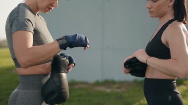 Açık havada spor giyimli iki kadın, birbirlerine kenetleniyorlar. Birbirlerine boks eldivenleri giydiriyorlar, takım çalışması yapıyorlar ve spor yapmaya hazırlanıyorlar..