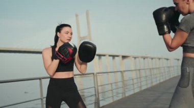 İki atletik kadın açık havada boks antrenmanına katılıyor, güneşli bir günde arka planda bir köprü ile yumruk pratiği yapıyorlar..