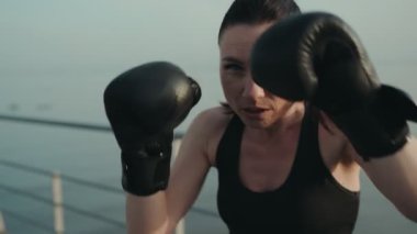 Kararlı atletik kadın deniz kenarındaki eldivenlerle boks antrenmanı yapıyor, gücünü ve dikkatini gösteriyor..