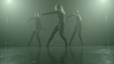 Üç dansçı stüdyoda ısınma hareketleri yapıyor, parlak ışıklara karşı siluetlenmiş, yüksek topuklu ayakkabılar giyiyor..
