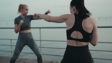 Odaklanmış kadın sporcular, sakin bir rıhtım kenarında boks antrenmanına çıkarak güç ve takım çalışması sergiliyorlar..