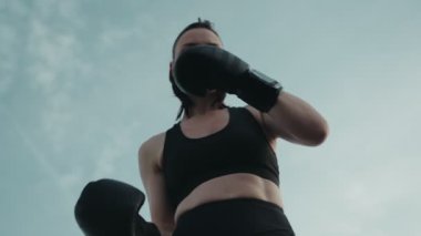 Açık gökyüzüne yumruk atan bir kadın boksörün düşük açılı çekimi. Güç, odaklanma ve kararlılık yayıyor..