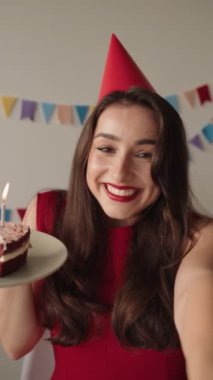 Parti şapkalı neşeli bir kadın, yanan bir doğum günü pastası ile bir selfie videosu çeker ve kapalı alanda kutlama anı yakalar..