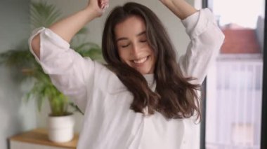 Neşeli ve çekici genç esmer kadın evde tek başına dans ediyor, parlak bir gülümsemeyle kamerayla meşgul..