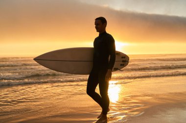 Gün batımında sahilde sörf tahtası taşıyan bir sörfçünün silueti sükunet ve macerayı yansıtıyor..