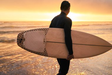 Sörf tahtasını elinde tutan bir sörfçü kıyıda duruyor, okyanusa bakıyor ve arkasında canlı bir günbatımı var..
