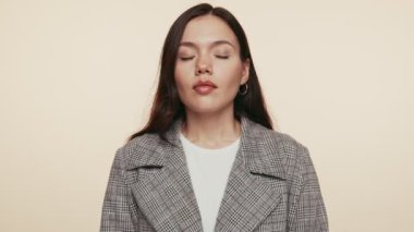 Sakin, genç bir kadın, gözleri kapalı, bej bir arka plan üzerinde derin bir nefes alıyor, dinginlik yayıyor ve şık bir kıyafetle dikkat çekmeye çalışıyor..