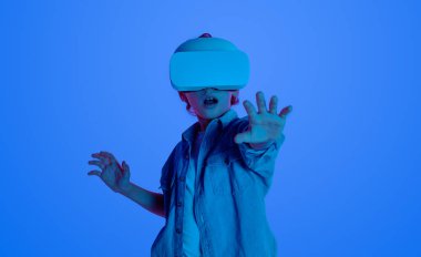 VR kulaklık takan çocuk, sanal çevreyle etkileşimde. Mavi arkaplan sanal gerçekliğin şaşırtıcı deneyimini vurgular.