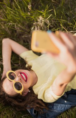 Sarı elbiseli ve güneş gözlüklü mutlu kadın çimlerin üzerinde güneşli havanın tadını çıkarırken selfie çekiyor..