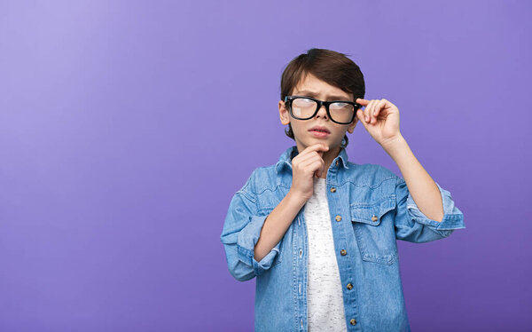 A young boy in glasses and a denim shirt looks thoughtful against a purple background. The image conveys curiosity and contemplation.