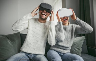 VR kulaklık takan ve modern bir oturma odasında eğlenen neşeli bir çift. Birlikte sanal gerçeklik deneyiminin tadını çıkarıyorlar..