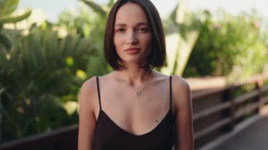 Siyah elbiseli kısa siyah saçlı genç bir kadın, kameraya hafifçe gülümsüyor. Tropikal bir bahçede duruyor..