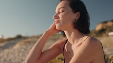 Genç bir kadın kumsalda huzur içinde oturur tenindeki güneşin sıcaklığının ve saçlarındaki hafif esintinin tadını çıkarır..