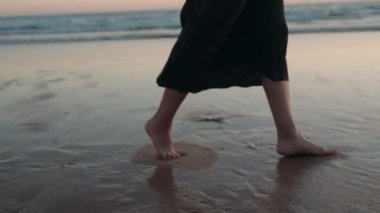 Siyah elbiseli bir kadın gün batımında kumsalın ıslak kumlarında yalınayak yürür. Ayakları kumda ayak izleri bırakıyor..