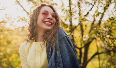 Neşeli kadın açık havada güneşli bir gün geçiriyor, şık güneş gözlükleri takıyor ve kot ceket giyiyor..