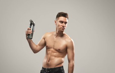 Tişörtsüz, zinde bir adam su şişesiyle rahatça poz veriyor. Sağlık, zindelik ve hidrasyon konsepti bir stüdyo ortamında yakalandı..