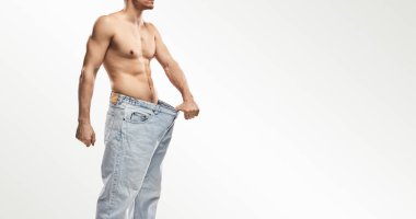Tişörtsüz bir adam kilosunu büyük beden kot pantolon giyerek gösteriyor. Spor başarısını ve vücut dönüşümünü simgeliyor..