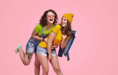 İki genç kadın birlikte eğleniyor, gülüyorlar ve pembe arka planda kaygısız bir anın tadını çıkarıyorlar..