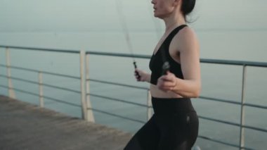 Odaklanmış bir kadın idman vitesi trenlerinde deniz kenarında bir gezinti güvertesinde ip atlıyor, vücut bulmuş zindelik ve kararlılık..