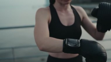 Odaklanmış atletik bir kadın açık havada boks antrenmanına katılıyor. Antrenman eldivenlerini, bulanık bir rıhtım arka planına takıyor..