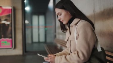 Düşünceli genç bir kadın bir tren istasyonunda oturuyor, akıllı telefonuna dalmış, sabırla şehir merkezine gitmesini bekliyor..