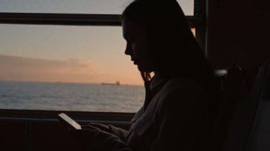 Gün batımında feribotta seyahat ederken akıllı telefonuyla emilmiş genç bir kadının silueti, sakinlik ve yalnızlık hissi uyandırıyor..