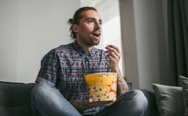 Genç adam kanepede oturuyor, yoğun bir film izliyor, patlamış mısır yiyor, odaklanma ve heyecan gösteriyor. Günlük ev ayarları.