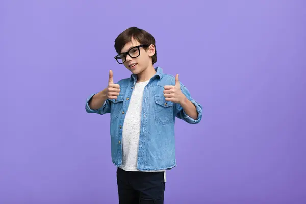 stock image A happy young boy wearing glasses and a denim jacket gives a double thumbs-up gesture, standing against a vibrant purple background.