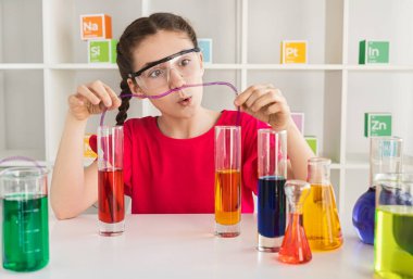 Meraklı genç kız güvenlik gözlüğü takıyor ve laboratuvar ortamında test tüpleriyle renkli bir kimya deneyi yapıyor..