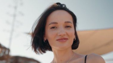 Kaygısız genç kadın kumsalda güneşli ve rüzgarlı bir günde kameraya gülümsüyor. Parmaklarını saçlarında gezdiriyor..