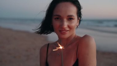 Gün batımında bir plajda tek başına bir maytapla gülümseyen ve kutlama yapan bir kadın. O mutlu ve sessiz güzelliğin tadını çıkarıyor..