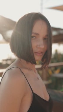 Kısa siyah saçlı ve siyah bluzlu güzel bir kadın güneşli bir günde kumsalda kameraya güvenle gülümsüyor..