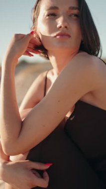 Genç bir kadın kumsalda oturuyor, teninde güneşin tadını çıkarıyor ve saçlarında hafif bir meltem esiyor..