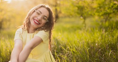 Gözlüklü neşeli bir kadın çimenli bir tarlada oturuyor, gün ışığında ışıl ışıl gülümsüyor. Mutluluğu, rahatlığı ve doğal güzelliği yakalar..