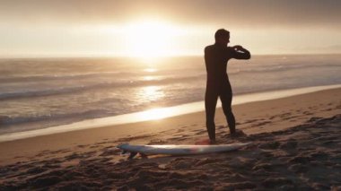 Bir sörfçü gün batımında sörf tahtasıyla kumsalda kollarını uzatır ve suya girmeye hazırlanır..