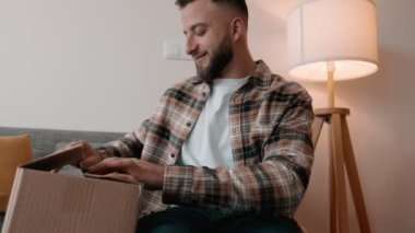 Gülümseyen sakallı, normal gömlekli bir adam evde bir karton kutuyu açıyor, siparişi görmek için sabırsızlanıyor ve seviniyor.