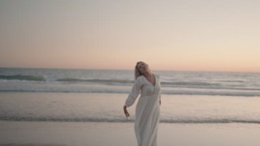 Beyaz elbiseli tasasız kadın gün batımında sahilde dans ediyor. Kolları açık, ve o anın özgürlüğünü kucaklıyor..