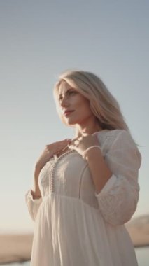Beyaz elbiseli neşeli, hamile bir kadın güneşli bir plaj gününün tadını çıkarıyor. Kameraya doğru yürürken gülümsüyor ve karnını tutuyor. Hamilelik, annelik, annelik ya da aile temaları için ideal.