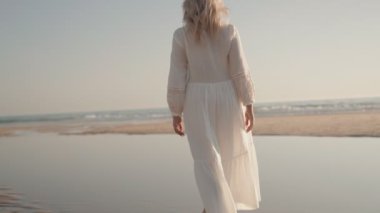 Beyaz elbiseli bir kadın kumlu bir sahilde yürüyor, okyanus dalgalarının dinginliğini ve güneşin sıcaklığını kucaklıyor..