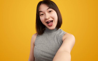 Neşeli genç bir kadın, canlı sarı bir arka plan önünde mutluluğunu ifade ederken eğlenceli bir selfie çekiyor. Keyif ve doğallığı yakalamak.